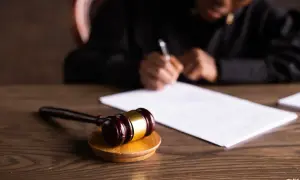Młotek sędziowski na drewnianym stole, w tle osoba pisząca na dokumencie.