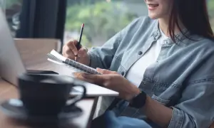 Kobieta pisze w notatniku, korzystając z prawa cytatu, by zanotować inspiracje. Obok kawa i laptop.