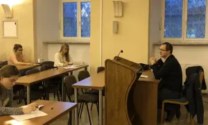 Referendarz sądowy prowadzi szkolenie. Kilka osób siedzi przy ławkach, słuchając i notując.