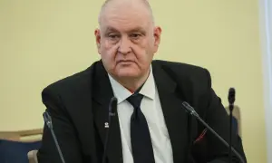 Prezes Trybunału Konstytucyjnego w garniturze, z przypinką z godłem Polski, siedzi przy stole z mikrofonami.