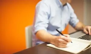Mężczyzna w niebieskiej koszuli pisze piórem w notatniku, pracując przy laptopie. Jego **prokura** jest widoczna w skupieniu na zadaniu.