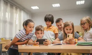 Grupa małoletnich uczniów skupiona wokół telefonu i tabletu, z zaciekawieniem oglądają coś na ekranach.