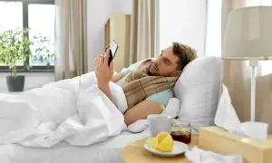 Chory mężczyzna leży w łóżku, przeglądając telefon. Obok kubek z herbatą, miód, cytryna i pudełko chusteczek. To jego siła wyższa, która usprawiedliwia nieobecność w pracy zgodnie z kodeksem pracy.