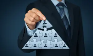 Biznesmen układa piramidę z puzzli, symbolizującą strukturę firmy. Każdy element to pracownik, a najwyższy to beneficjent, który czerpie korzyści.