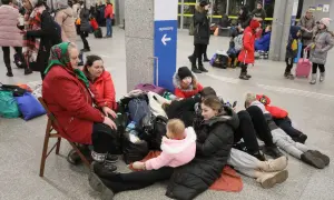 Rodzina z dziećmi, w tym niemowlę, siedzi na podłodze z bagażami, czekając na pomoc. Ich status uchodźcy jest widoczny w ich sytuacji.