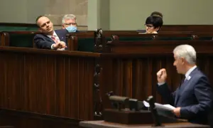 Posłowie w ławach sejmowych, jeden z nich z maską na twarzy, słuchają przemówienia. Atmosfera jak na trybunale stanu.