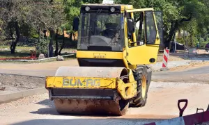 Żółty pojazd wolnobieżny, walec drogowy Bomag, pracuje na budowie drogi, ubijając nawierzchnię.