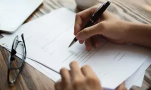 Dłoń z długopisem nad dokumentem, podpisując umowę. W tle okulary i tablet. Kluczowe jest porozumienie stron.