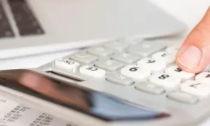 Dłoń naciska klawisze kalkulatora obok laptopa i dokumentów.