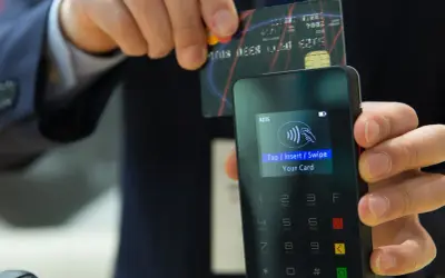 Zbliżenie na terminal płatniczy z monitorem pokazującym instrukcję "Tap/Insert/Swipe Your Card". Karta zbliżeniowa jest trzymana nad czytnikiem.