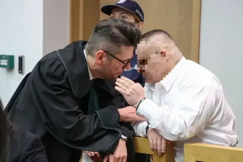 Prawnik pochyla się nad oskarżonym, omawiając, czy oskarżony może zobaczyć zeznania świadków.