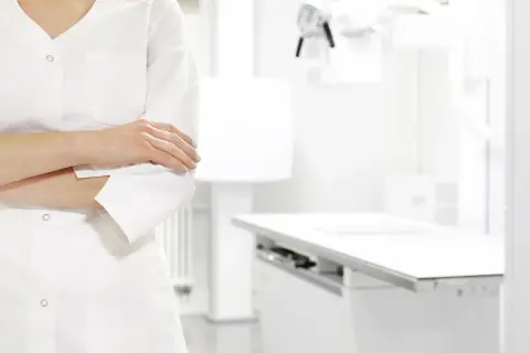 Lekarz w białym kitlu, z rękami skrzyżowanymi na piersi, w tle gabinetu zabiegowego. Zastanawia się nad ubezpieczeniem zdrowotnym ZUS dla niepracujących.