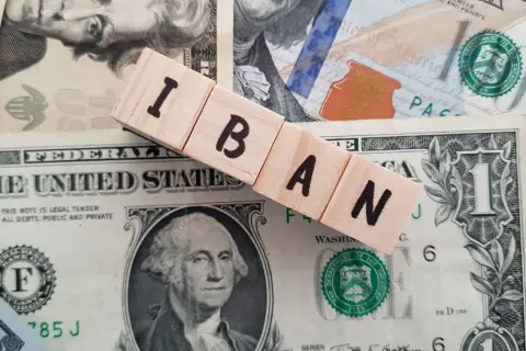 Drewniane klocki z literami IBAN leżą na banknotach dolarowych. IBAN to międzynarodowy numer rachunku bankowego.