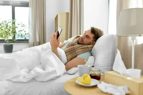 Chory mężczyzna leży w łóżku, przeglądając telefon. Obok kubek z herbatą, miód, cytryna i pudełko chusteczek. To jego siła wyższa, która usprawiedliwia nieobecność w pracy zgodnie z kodeksem pracy.