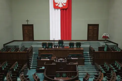 Pusta sala sejmowa, gotowa na głosowanie weta prezydenta. Biało-czerwona flaga i godło Polski na ścianie.