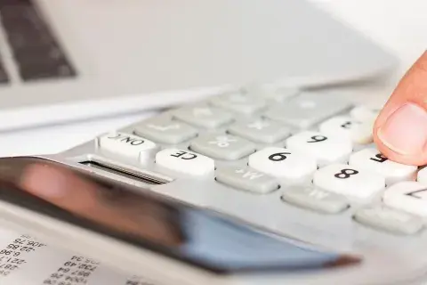Dłoń naciska klawisze kalkulatora obok laptopa i dokumentów.