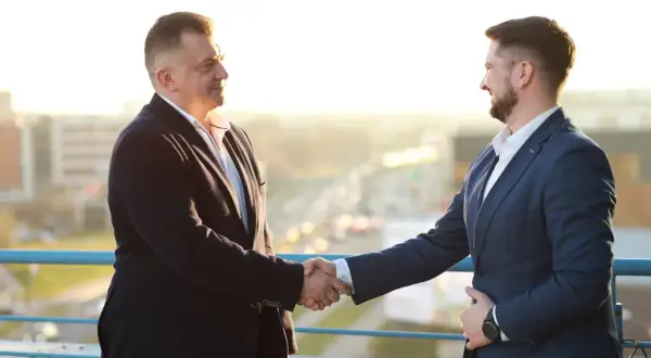Dwóch mężczyzn w garniturach ściska sobie dłonie na tle miasta.