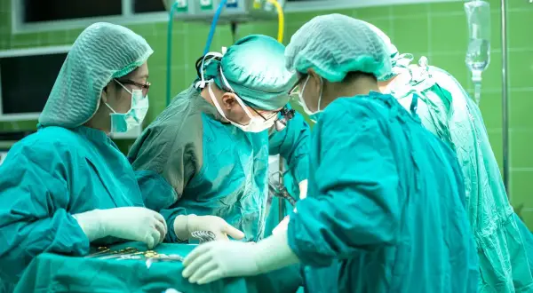 Zespół chirurgów w sterylnych strojach pracuje przy stole operacyjnym. W tle widać aparaturę medyczną. Sytuacja budzi niepokój, czy nieumyślne spowodowanie śmierci nie jest tu możliwe.