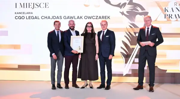 Pięć osób w strojach formalnych stoi na scenie, odbierając nagrodę.