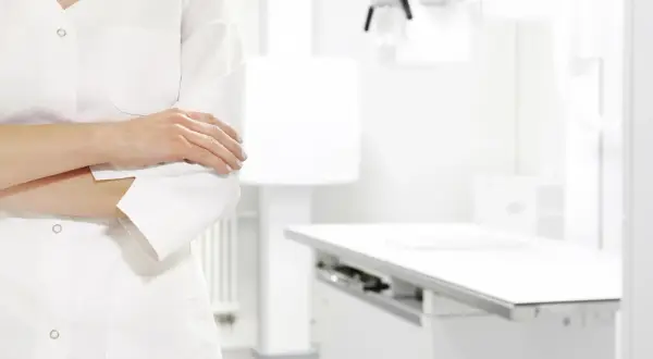 Lekarz w białym kitlu, z rękami skrzyżowanymi na piersi, w tle gabinetu zabiegowego. Zastanawia się nad ubezpieczeniem zdrowotnym ZUS dla niepracujących.