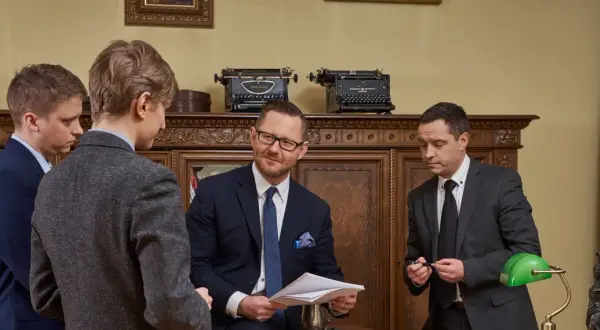 Czterech mężczyzn w garniturach w gabinecie, dyskutujących nad dokumentami.