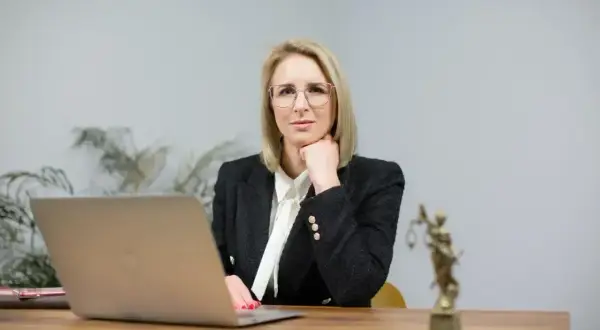 Kobieta w okularach i żakiecie siedzi przy biurku z laptopem i statuetką Temidy.
