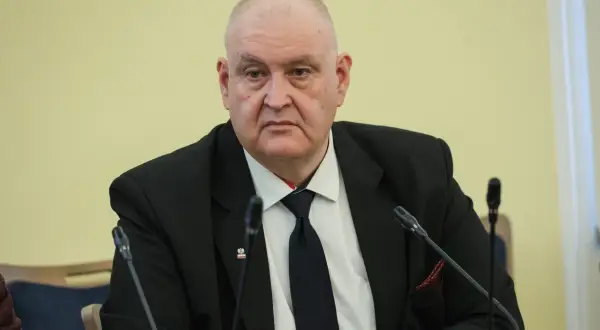 Prezes Trybunału Konstytucyjnego w garniturze, z przypinką z godłem Polski, siedzi przy stole z mikrofonami.