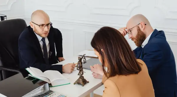Prawnik omawia z klientami, co grozi za niewykonanie prawomocnego wyroku sądu. Na biurku dokumenty i Statua Temidy.