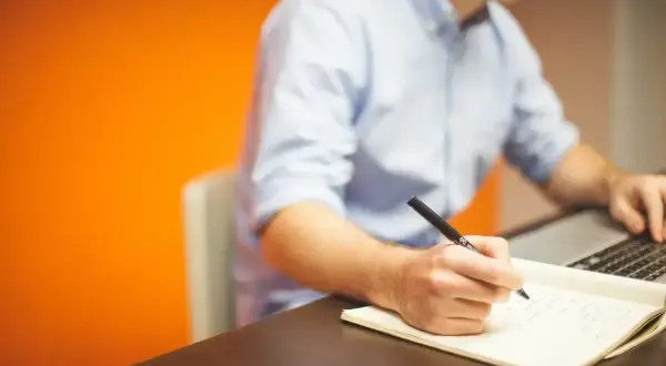 Mężczyzna w niebieskiej koszuli pisze piórem w notatniku, pracując przy laptopie. Jego **prokura** jest widoczna w skupieniu na zadaniu.