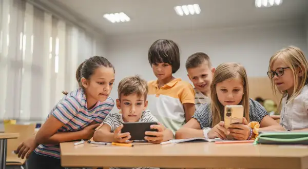 Grupa małoletnich uczniów skupiona wokół telefonu i tabletu, z zaciekawieniem oglądają coś na ekranach.
