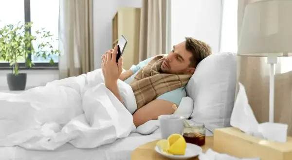 Chory mężczyzna leży w łóżku, przeglądając telefon. Obok kubek z herbatą, miód, cytryna i pudełko chusteczek. To jego siła wyższa, która usprawiedliwia nieobecność w pracy zgodnie z kodeksem pracy.