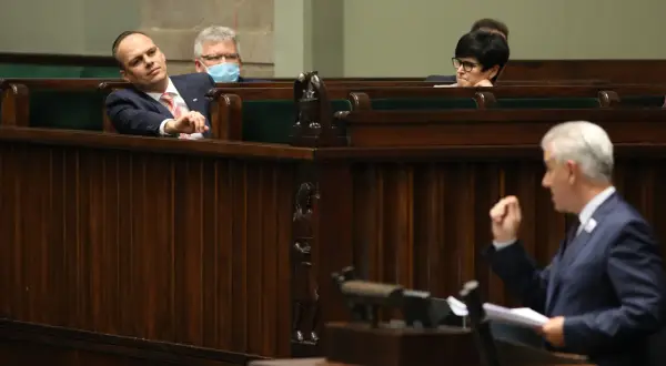 Posłowie w ławach sejmowych, jeden z nich z maską na twarzy, słuchają przemówienia. Atmosfera jak na trybunale stanu.