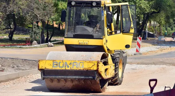 Żółty pojazd wolnobieżny, walec drogowy Bomag, pracuje na budowie drogi, ubijając nawierzchnię.