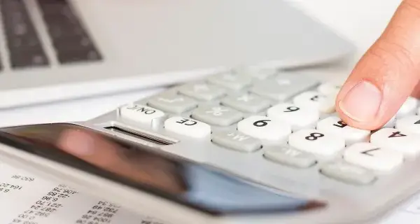 Dłoń naciska klawisze kalkulatora obok laptopa i dokumentów.