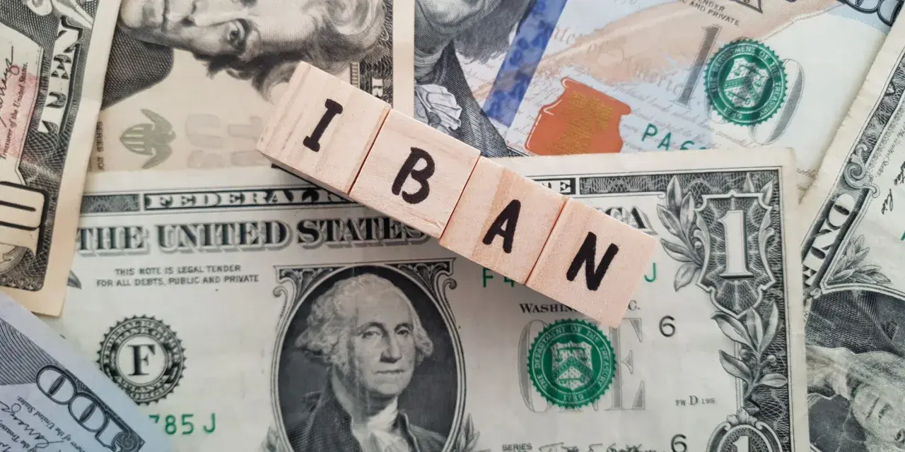Drewniane klocki z literami IBAN leżą na banknotach dolarowych. IBAN to międzynarodowy numer rachunku bankowego.