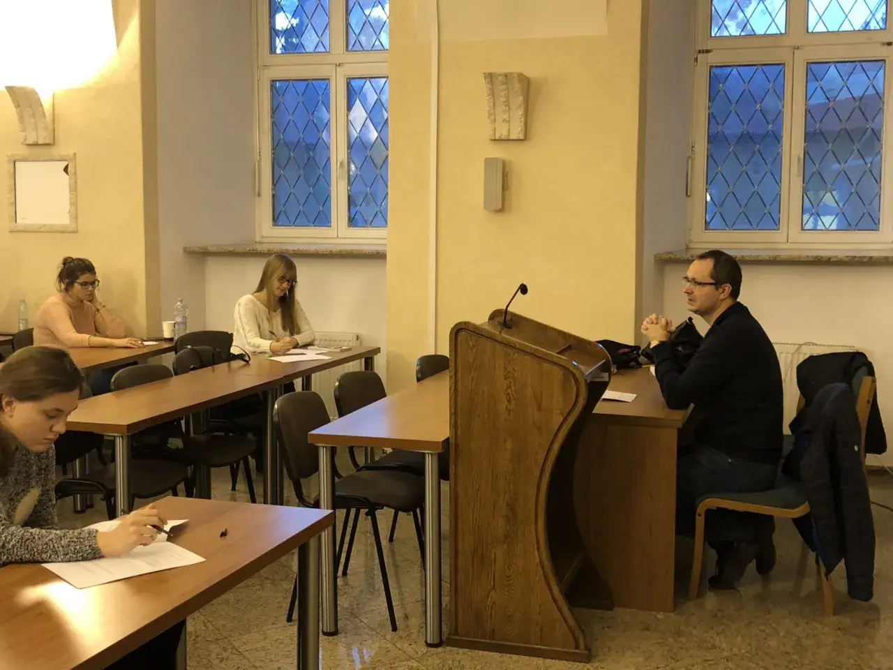 Referendarz sądowy prowadzi szkolenie. Kilka osób siedzi przy ławkach, słuchając i notując.