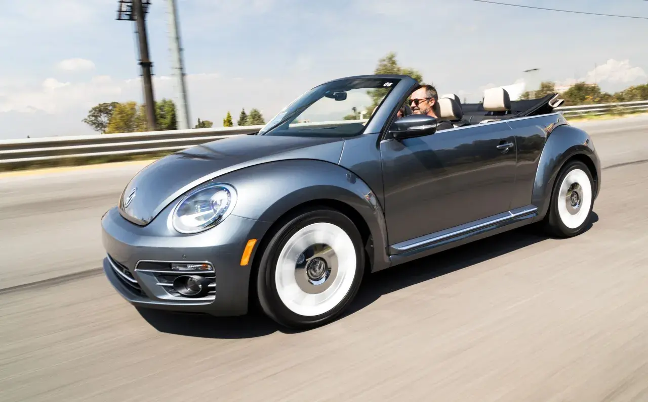 Szary kabriolet Volkswagen Beetle jedzie po drodze. Mężczyzna w okularach przeciwsłonecznych prowadzi auto. To idealny pojazd na letnie wycieczki, jak ta opisana w umowie użyczenia samochodu.