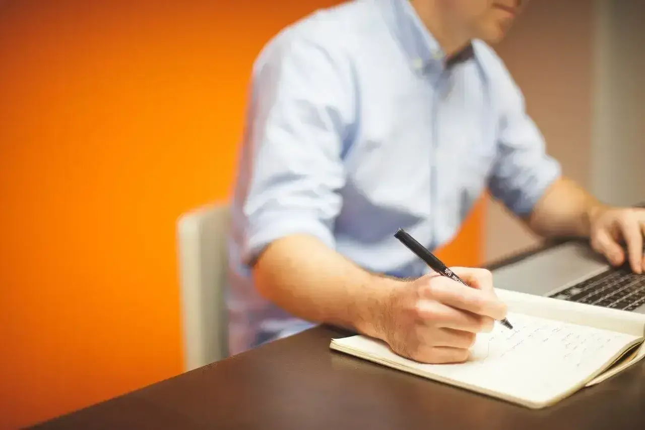 Mężczyzna w niebieskiej koszuli pisze piórem w notatniku, pracując przy laptopie. Jego **prokura** jest widoczna w skupieniu na zadaniu.
