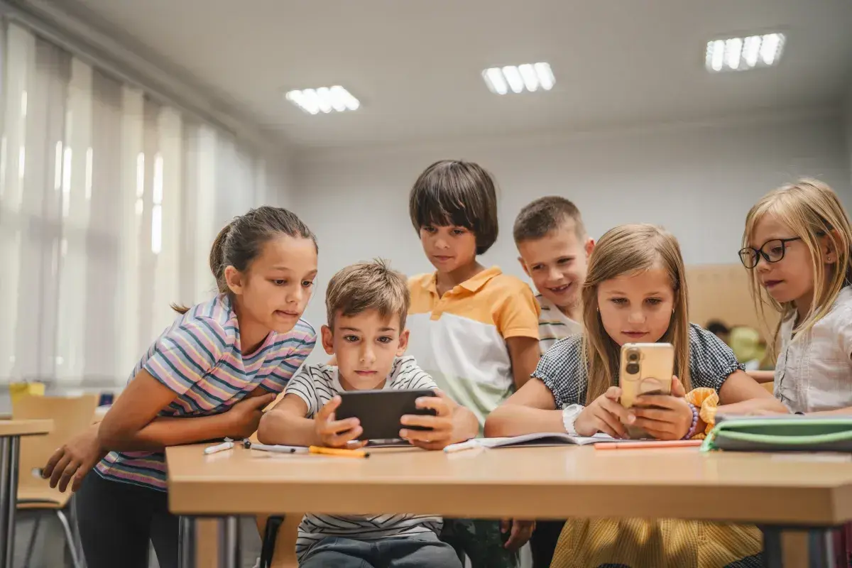 Grupa małoletnich uczniów skupiona wokół telefonu i tabletu, z zaciekawieniem oglądają coś na ekranach.