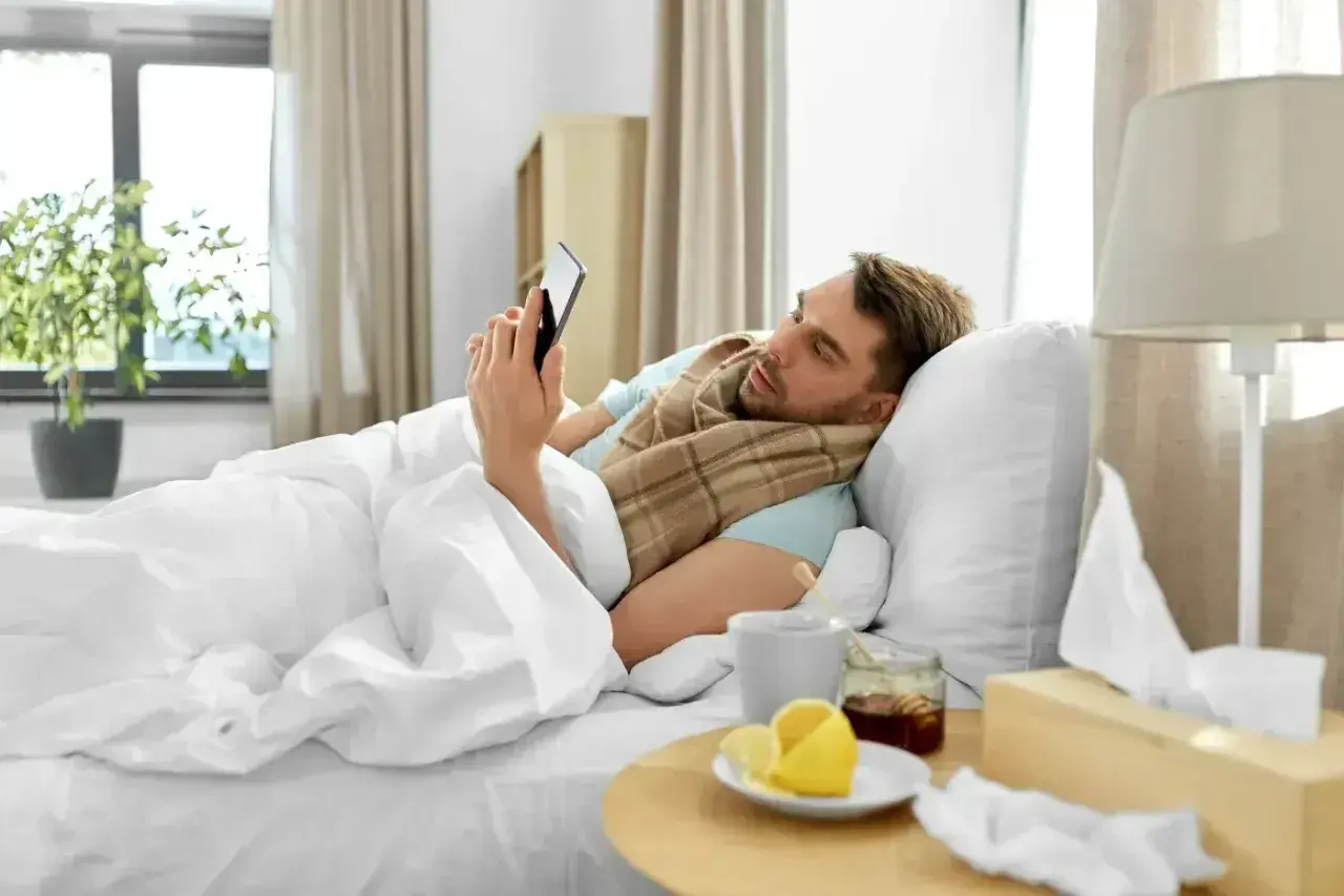 Chory mężczyzna leży w łóżku, przeglądając telefon. Obok kubek z herbatą, miód, cytryna i pudełko chusteczek. To jego siła wyższa, która usprawiedliwia nieobecność w pracy zgodnie z kodeksem pracy.
