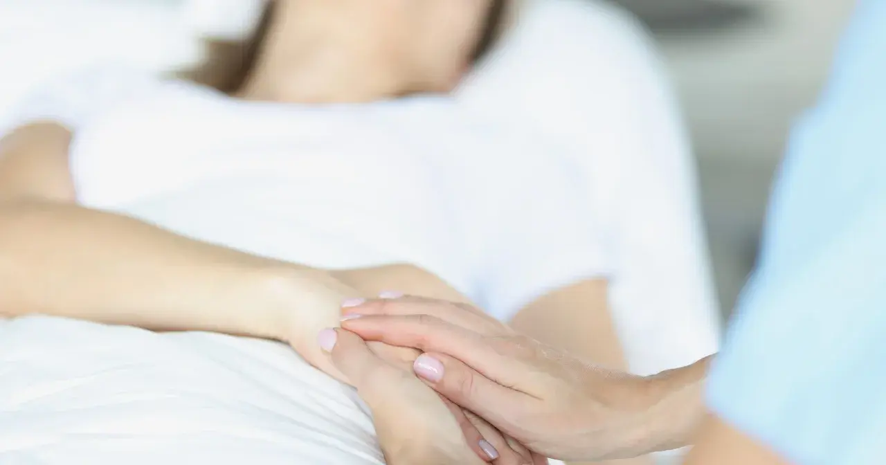 Dwie dłonie splatają się na białym tle. Jedna dłoń, należąca do pacjenta, jest blada. Druga, należąca do opiekuna, delikatnie ją obejmuje, symbolizując wsparcie w trudnych chwilach, gdy rozważane jest co to eutanazji.
