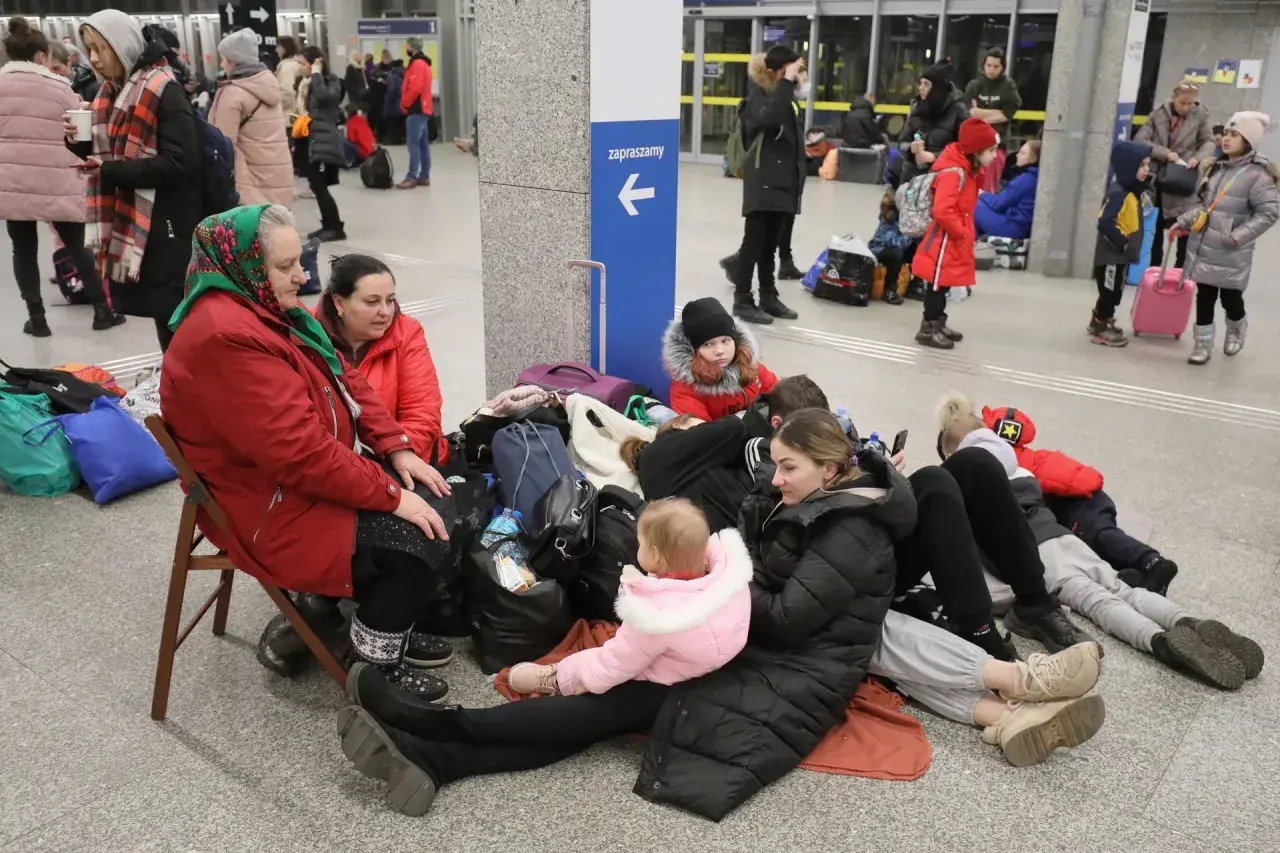 Rodzina z dziećmi, w tym niemowlę, siedzi na podłodze z bagażami, czekając na pomoc. Ich status uchodźcy jest widoczny w ich sytuacji.