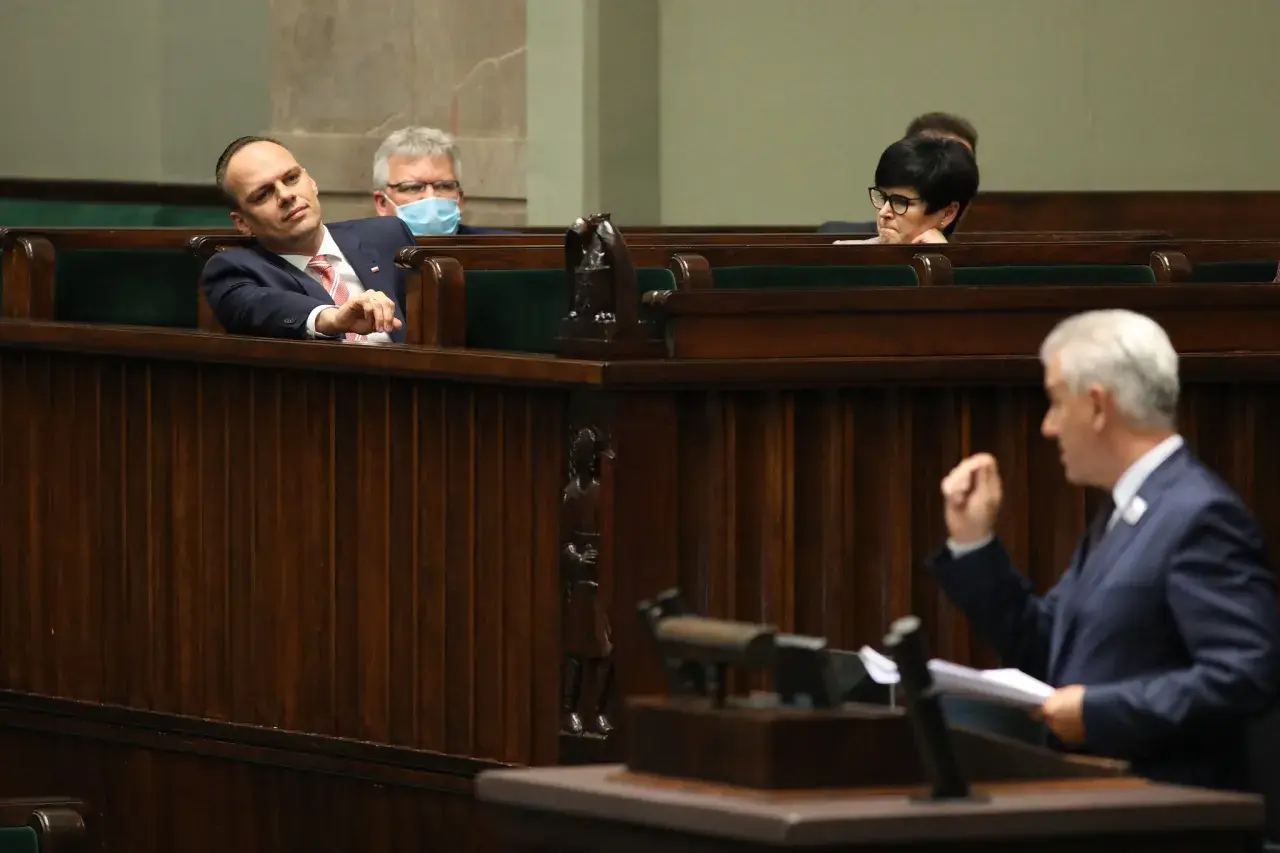 Posłowie w ławach sejmowych, jeden z nich z maską na twarzy, słuchają przemówienia. Atmosfera jak na trybunale stanu.