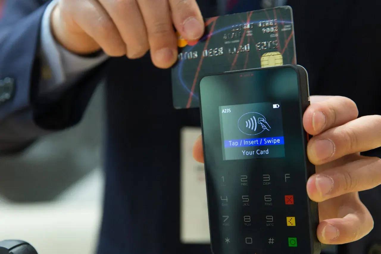 Zbliżenie na terminal płatniczy z monitorem pokazującym instrukcję "Tap/Insert/Swipe Your Card". Karta zbliżeniowa jest trzymana nad czytnikiem.