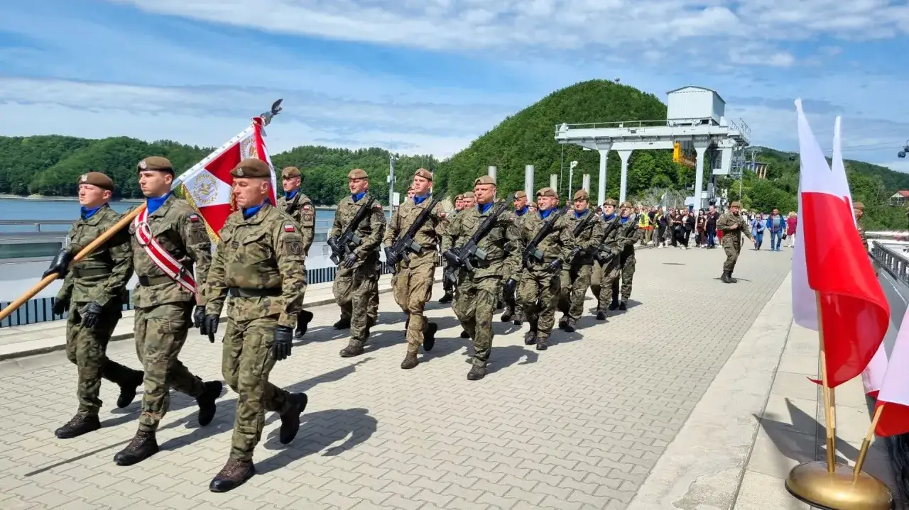 Żołnierze w pełnym rynsztunku maszerują z flagą, składając przysięgę w malowniczej scenerii nad jeziorem.