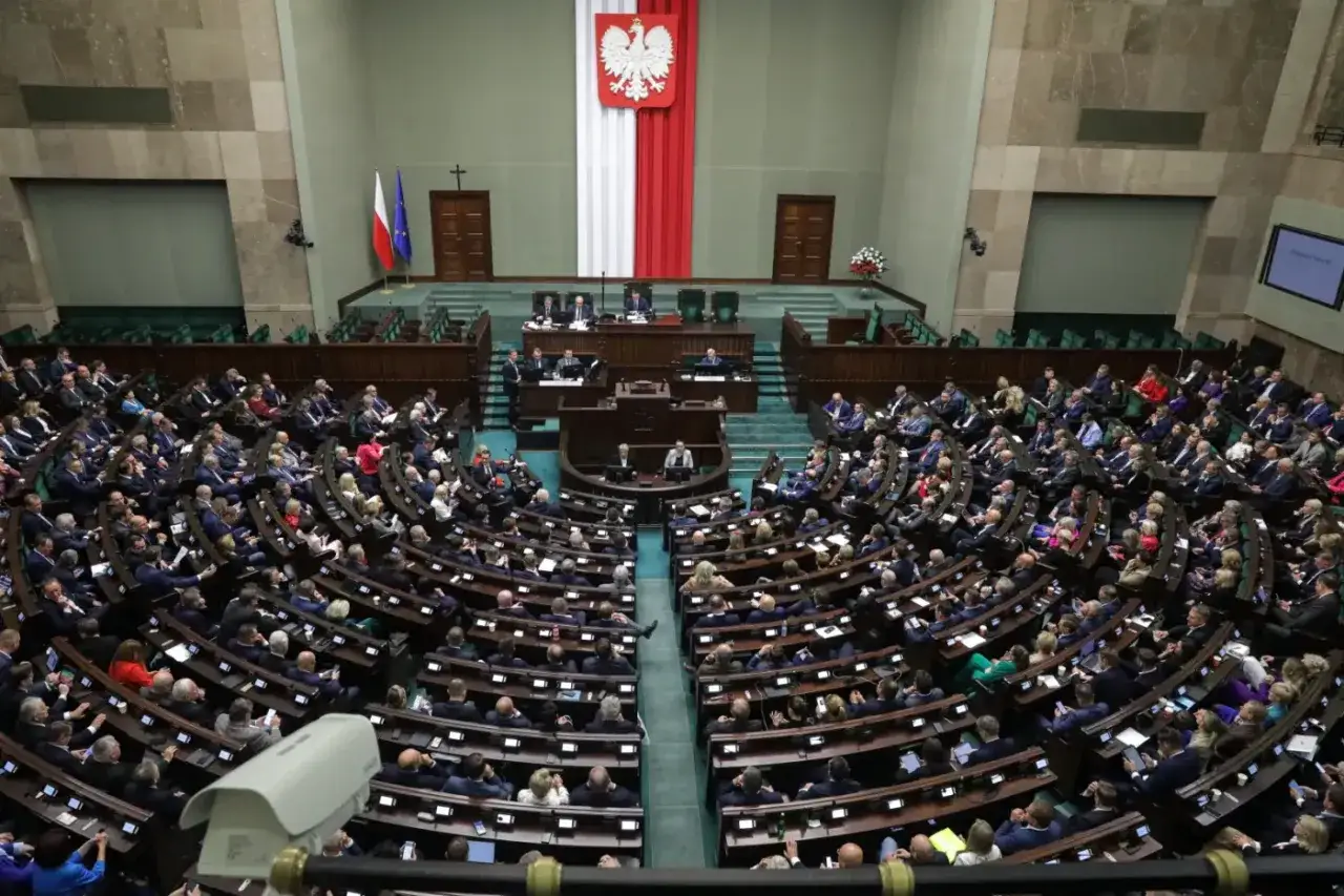 Posiedzenie Sejmu, gdzie trwa obstrukcja parlamentarna. Widać wielu posłów siedzących w ławach, flagi Polski i UE, a także godło państwowe.