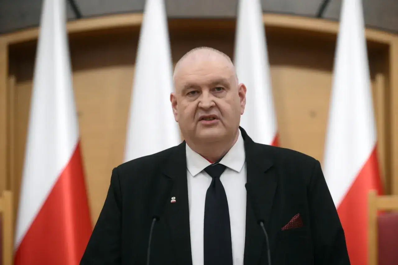 Prezes Trybunału Konstytucyjnego w garniturze, z polskimi flagami w tle.