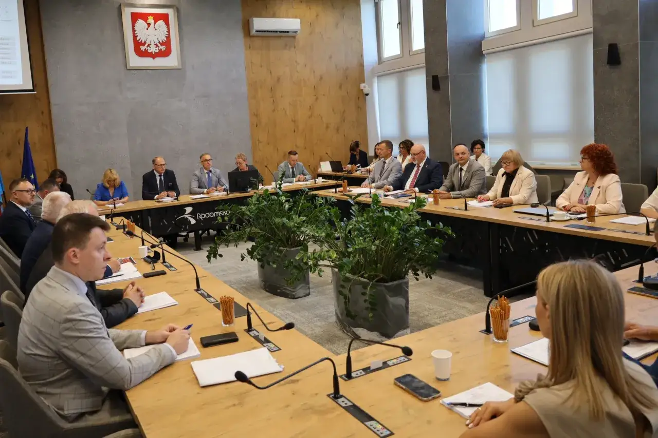 Obrady w sali konferencyjnej, dyskusja o tym, absolutorium co to. Ludzie siedzą przy stole, dokumenty, mikrofony.