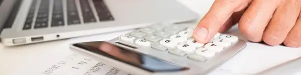 Dłoń naciska klawisze kalkulatora obok laptopa i dokumentów.