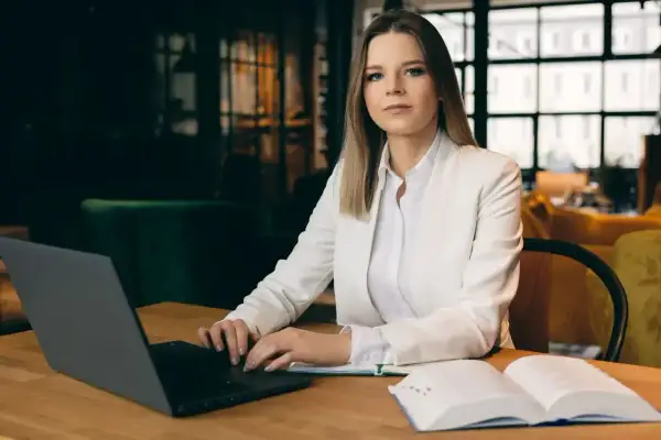 Kobieta w białej marynarce przy laptopie i otwartej księdze. Zastanawia się, jak zwracać się do adwokata, by uzyskać profesjonalną pomoc.